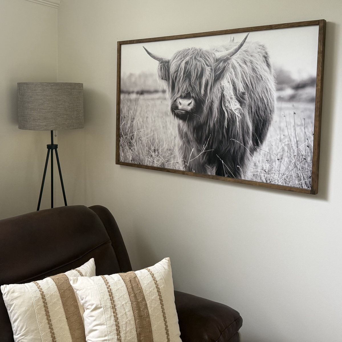 A corner of the living room: a brown leather sofa with cream striped pillows, a tripod floor lamp, and a large black-and-white print of a Highland cow in a wood frame.