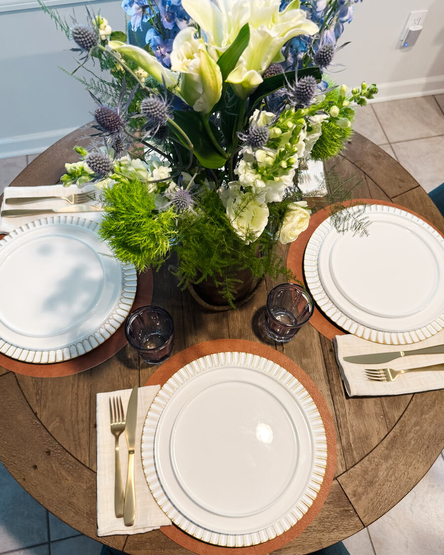A round table set for three, white plates on wood chargers, a low arrangement of lilies and delphinium.