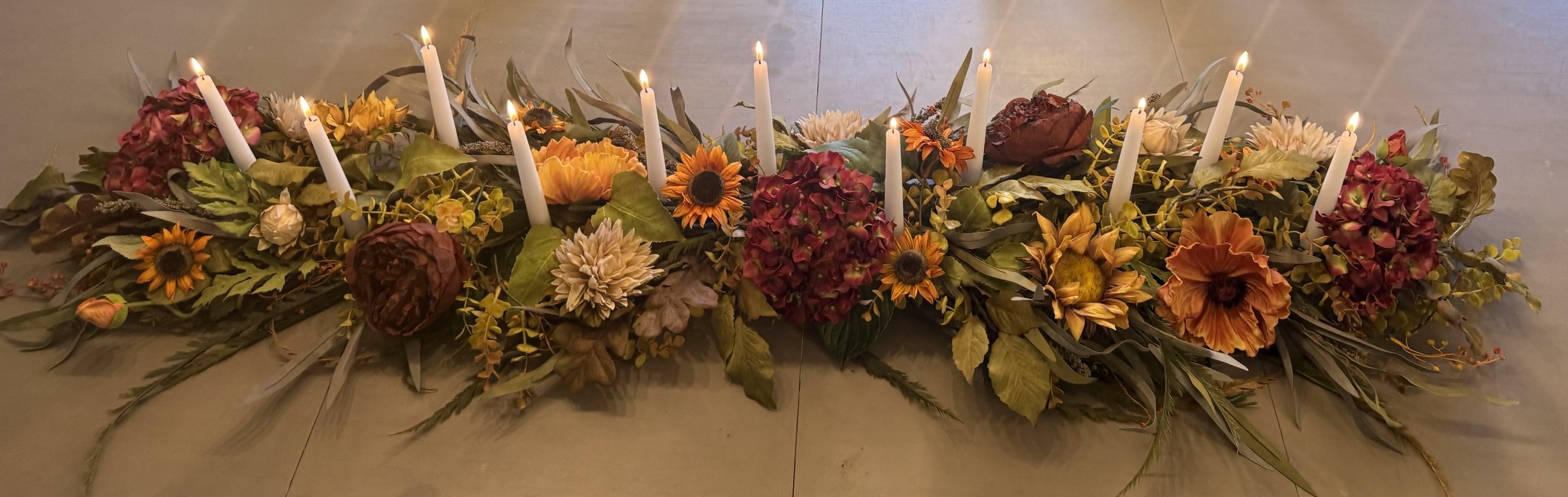A long autumn runner of burgundy hydrangea, rust and gold sunflowers, cream dahlias and greenery on a pale wood table, a row of lit white tapers rising out of it.