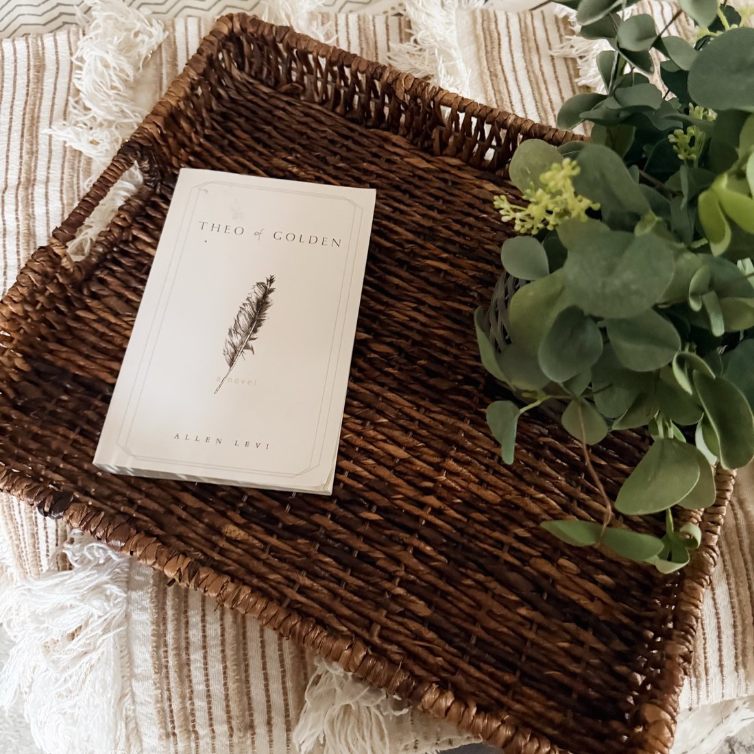 The novel Theo of Golden by Allen Levi in a woven basket on a fringed throw, with eucalyptus.