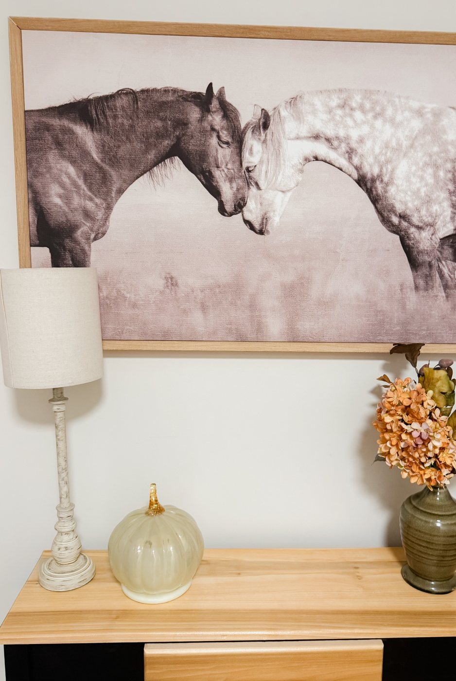 A console table under a framed print of two horses, with a linen lamp, a glass pumpkin candle and a green vase of autumn hydrangea.