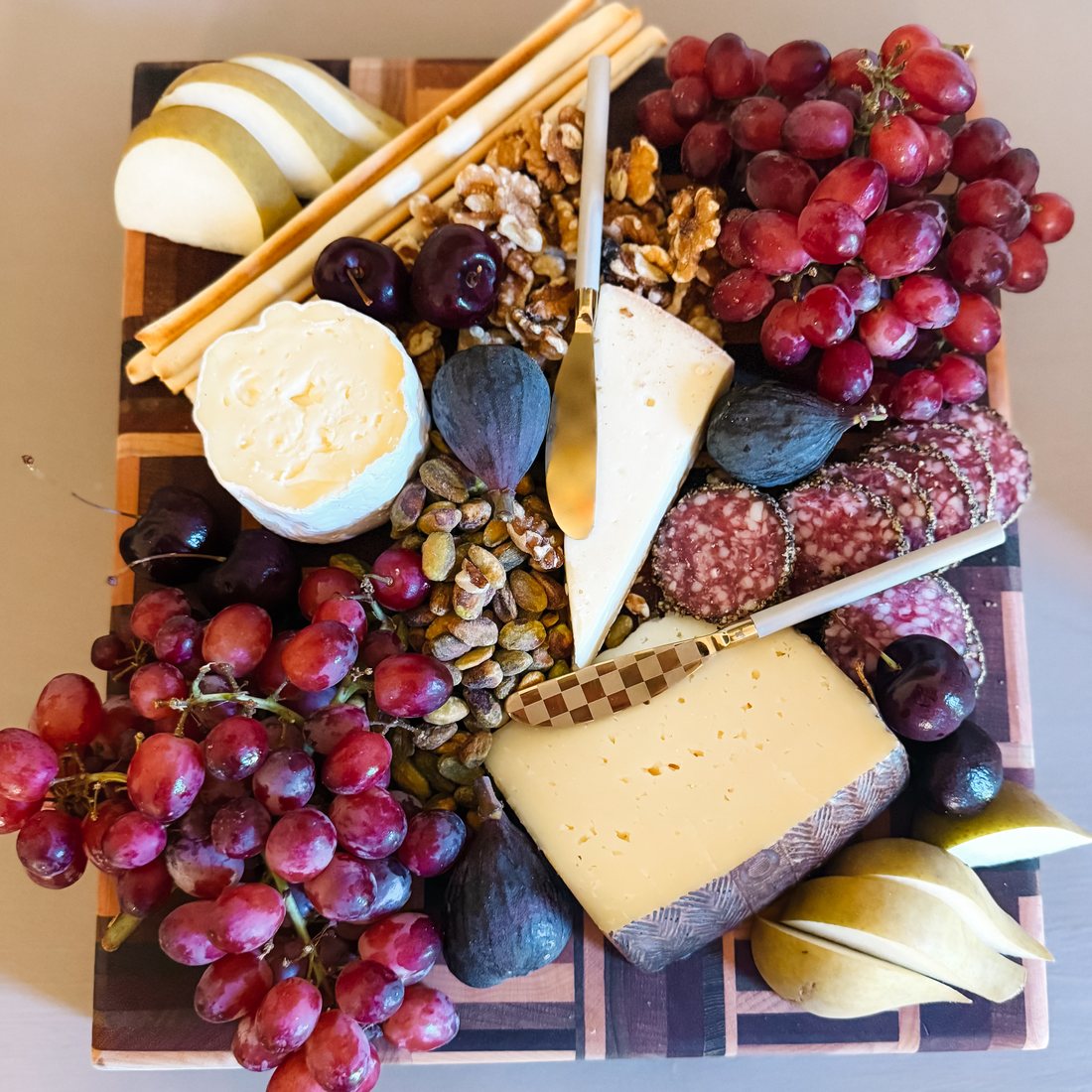 An autumn cheese board with figs, grapes, pears, salami and walnuts, a checked-handle cheese knife and a gold spreader resting on it.