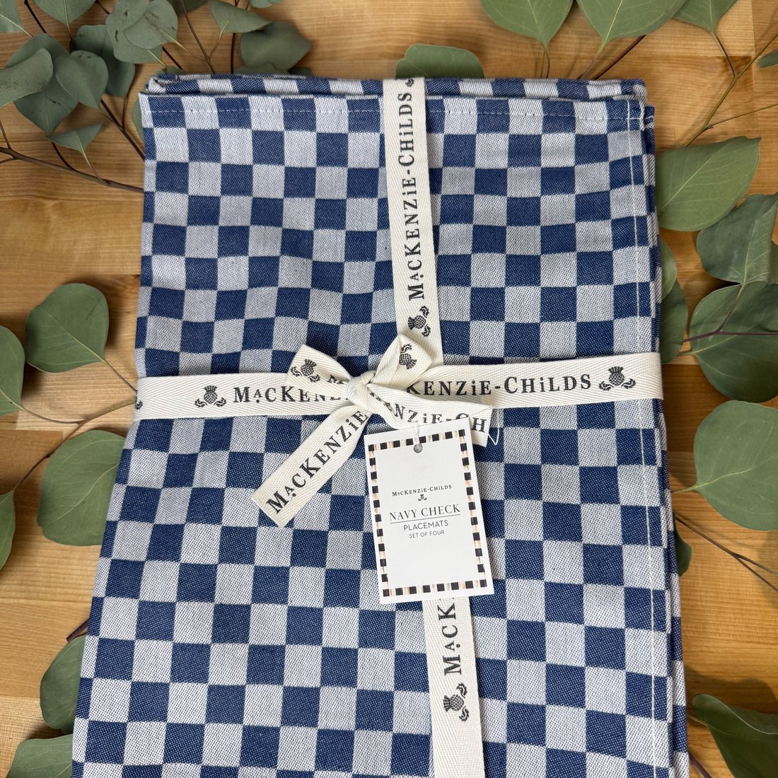 MacKenzie-Childs Navy Check placemats, tied with the maker&rsquo;s ribbon, on a wooden table with eucalyptus.
