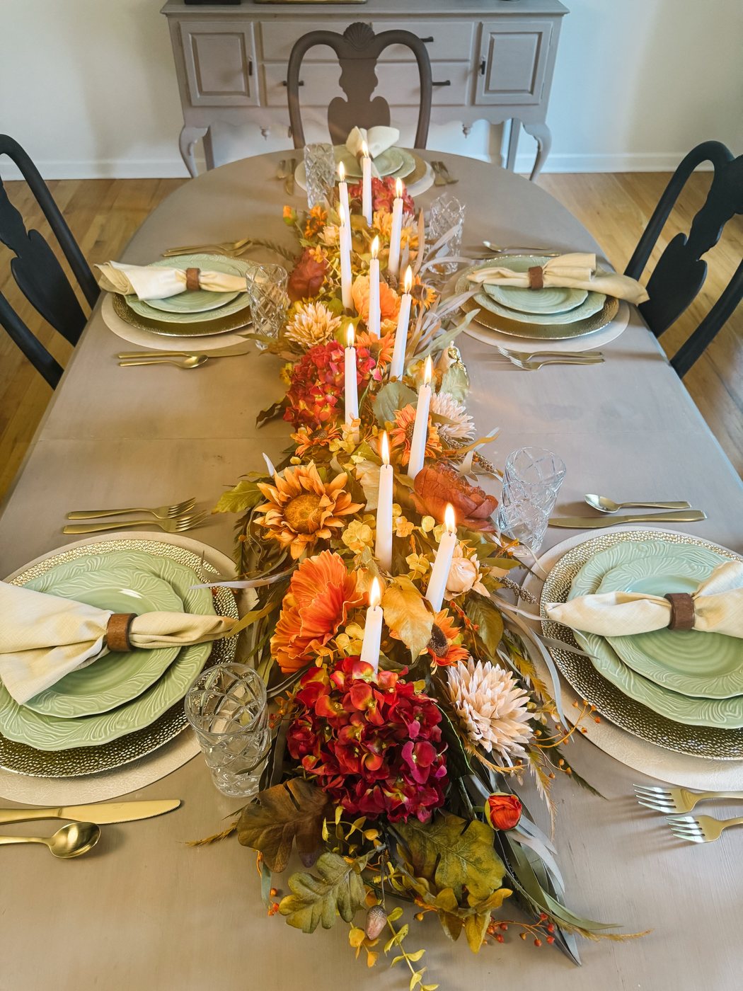 Looking down a long table set for autumn: green leaf-pattern plates on hammered gold chargers and cream placemats, linen napkins in leather rings, gold flatware, crystal glasses, and a runner of sunflowers, hydrangea and lit white tapers down the middle.