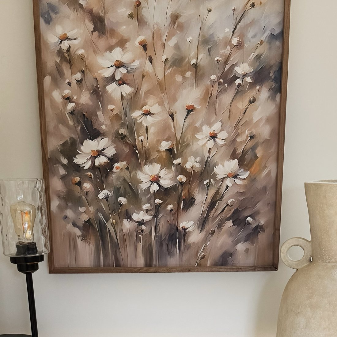 A framed painting of loose white wildflowers in blush, taupe and slate, hung above a marble console between a glass lamp and a cream stone vase.