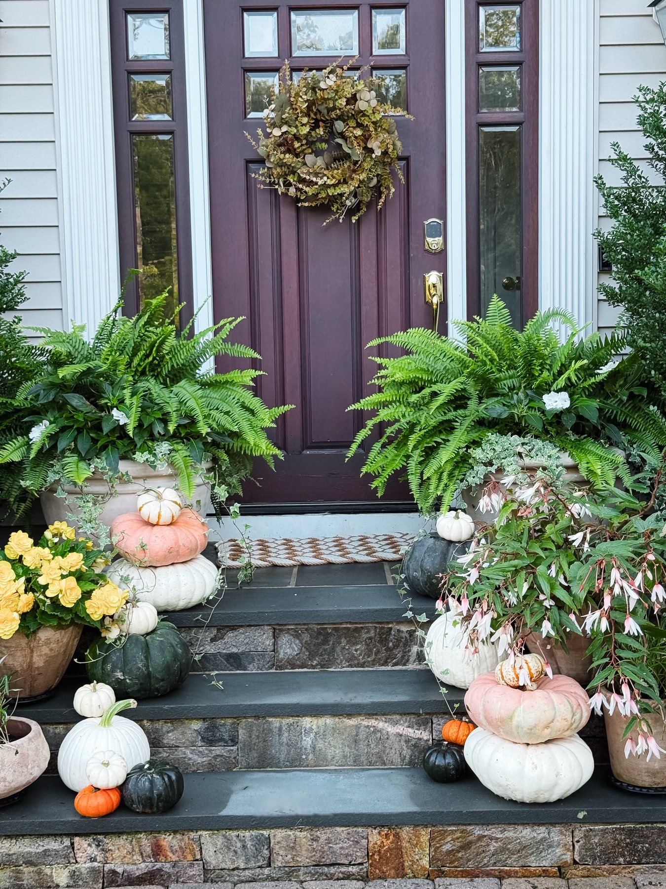 The front steps in autumn: a plum front door with a eucalyptus wreath, ferns in stone urns on either side, and heirloom pumpkins in pink, white and slate stacked down the bluestone steps beside yellow and pink begonias.