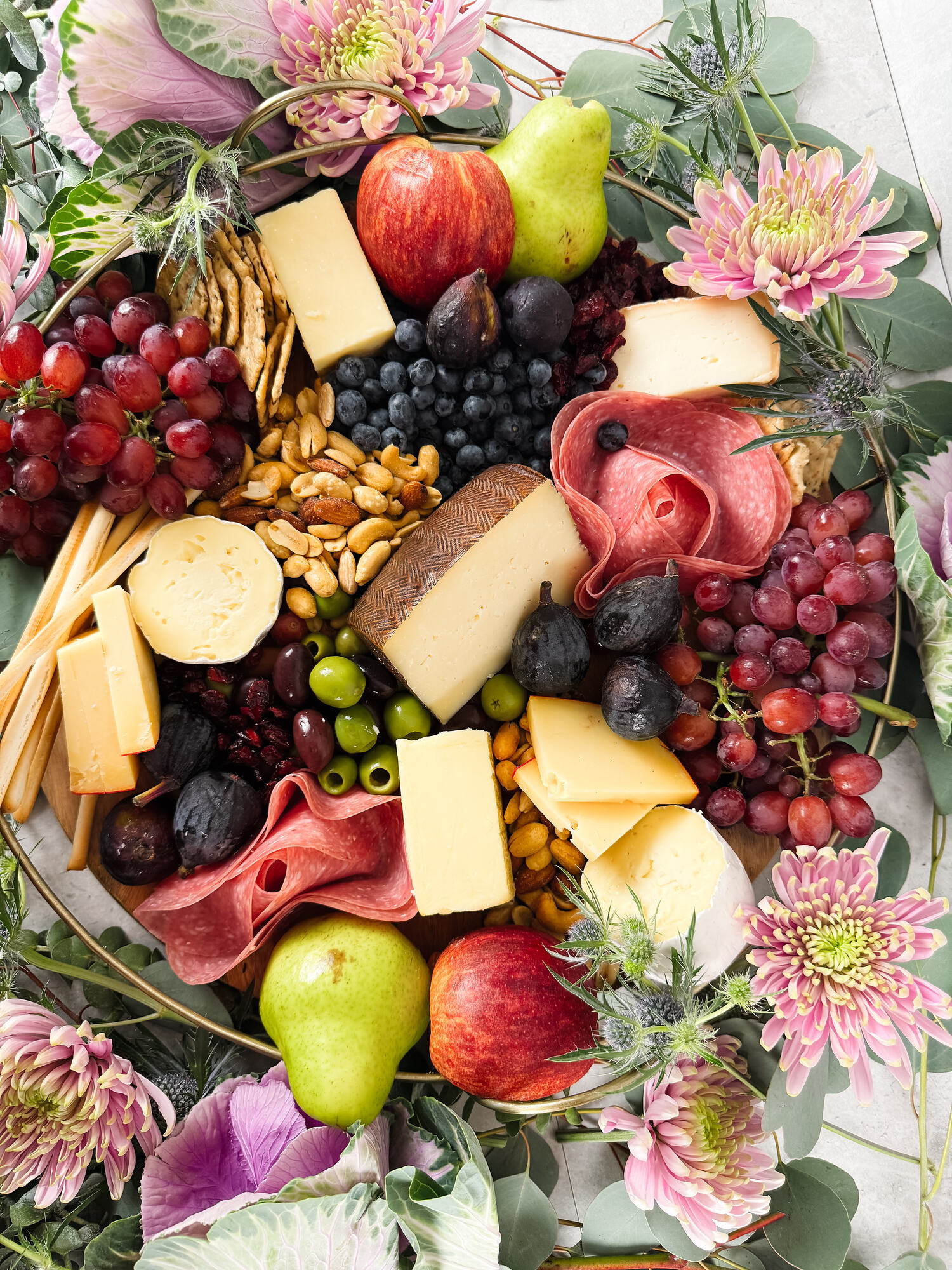 An overhead grazing board of cheeses, cured meats, figs, grapes and pears, ringed with eucalyptus and pink chrysanthemum on pale stone.