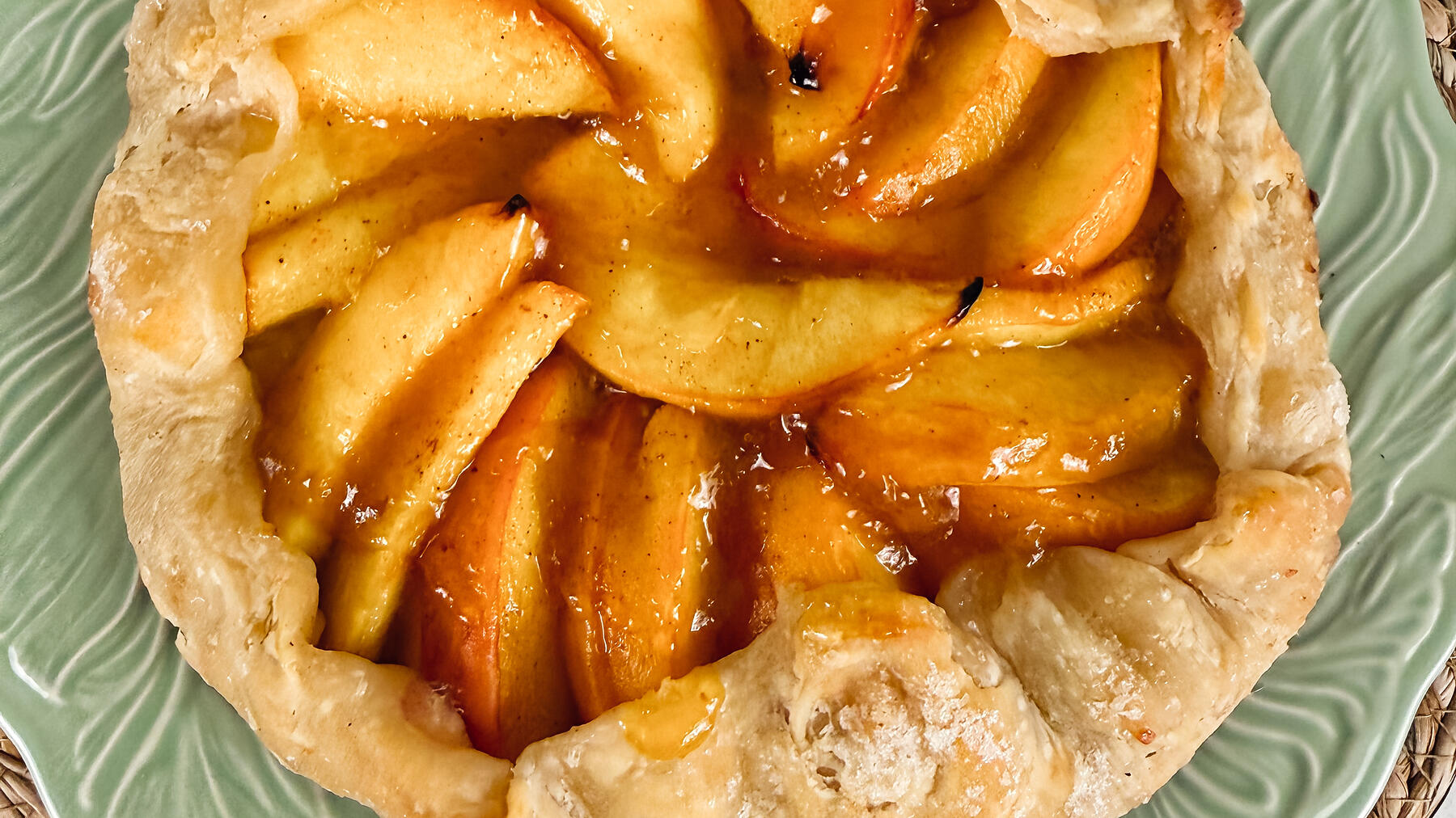 An overhead peach galette on a green leaf-pattern plate, the pastry folded loosely over sliced fruit, on pale stone.
