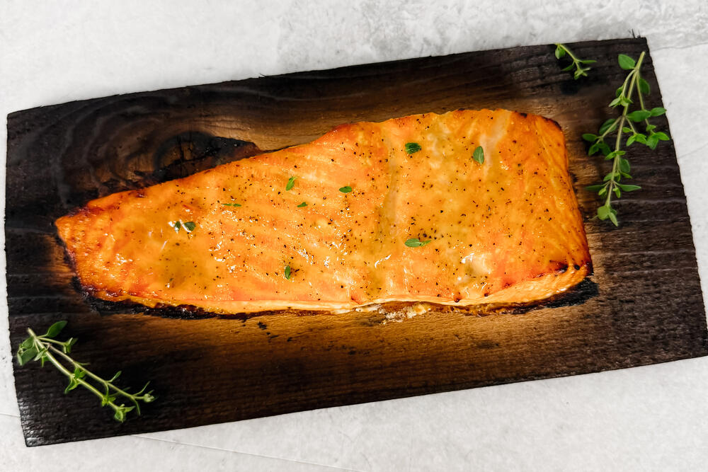 A roasted salmon fillet on a charred cedar plank with sprigs of thyme, on pale stone.