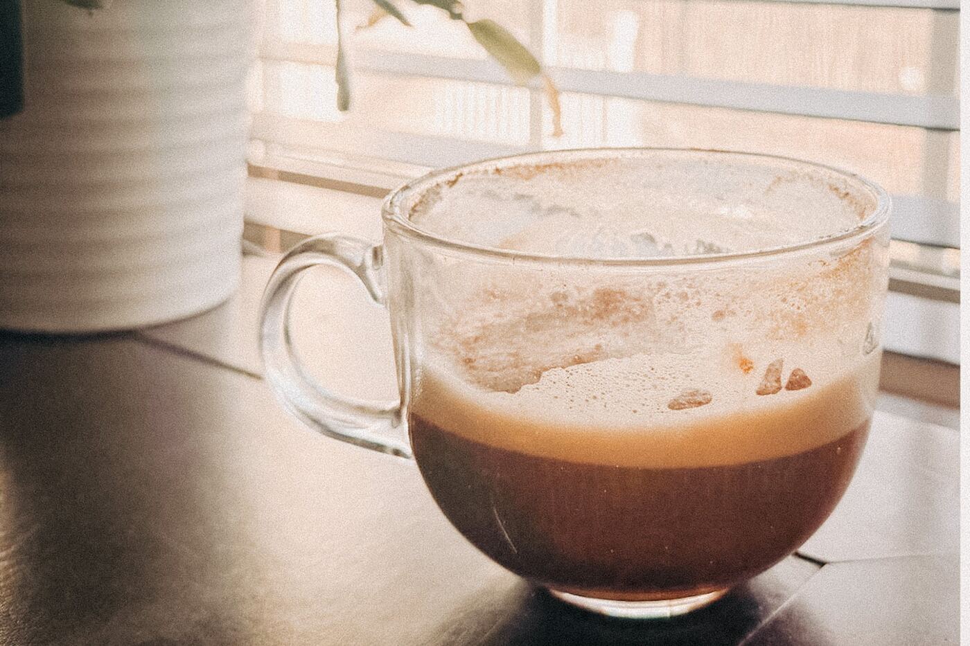 A glass mug of coffee with cinnamon on the counter by a bright window.