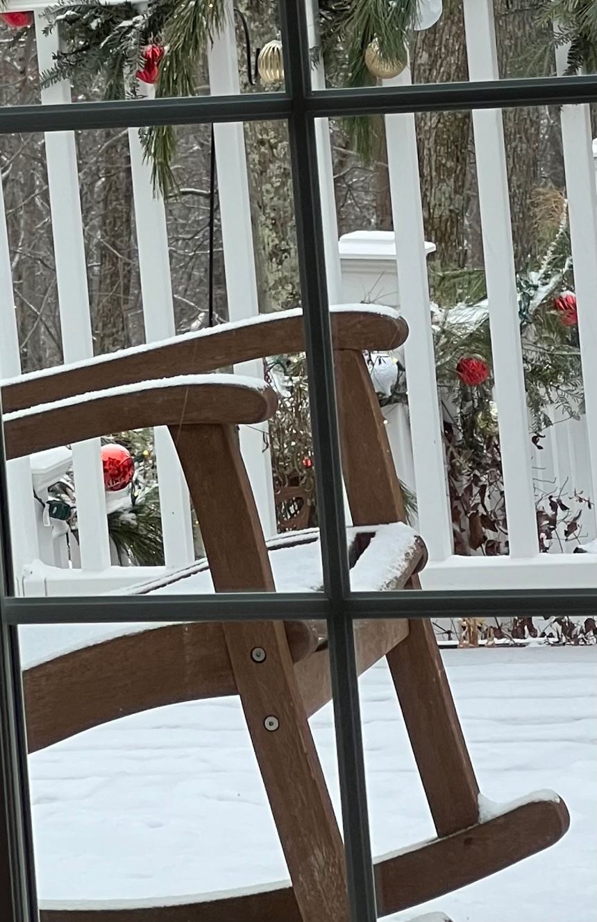 A wooden rocking chair on a snow-covered porch, seen through the window.