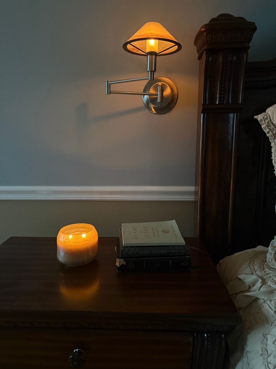 A lit candle and a stack of books on the nightstand, lamplight low.