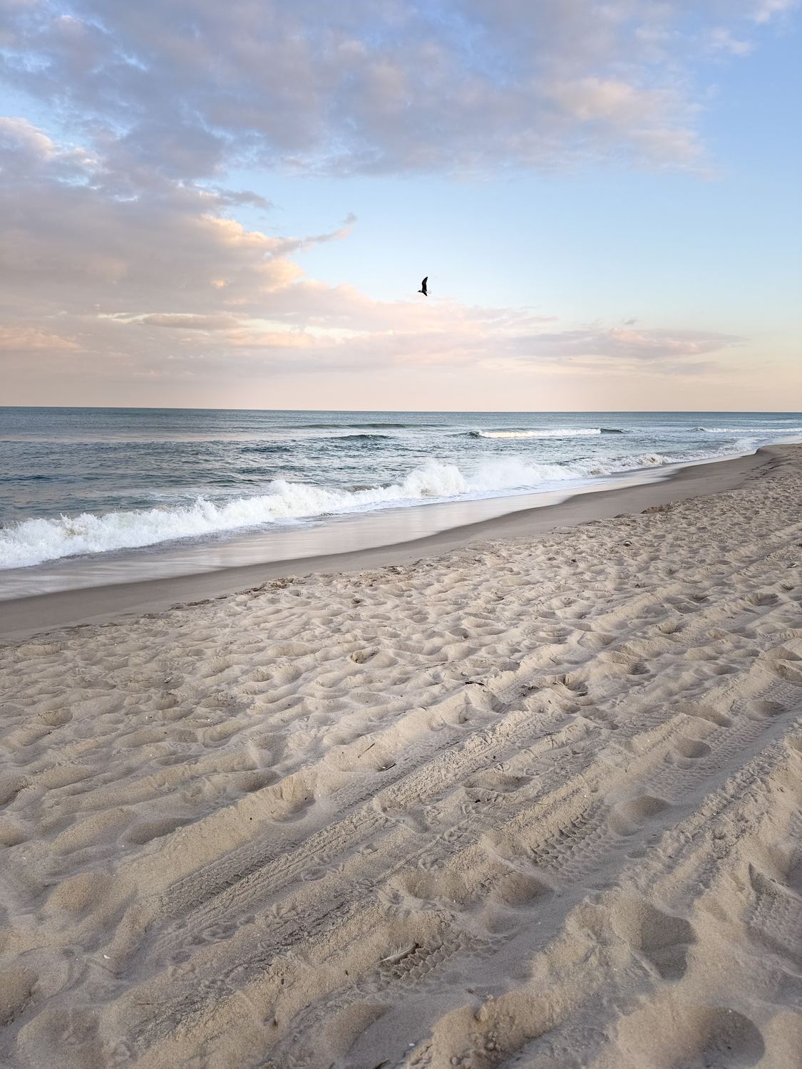 A wide, empty stretch of beach with footprints in the sand under a hard blue sky.