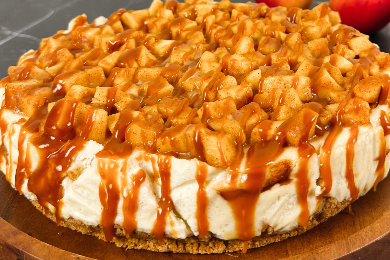 Caramel apple cheesecake on a wooden board, diced cinnamon apples piled on top and salted caramel running down the sides