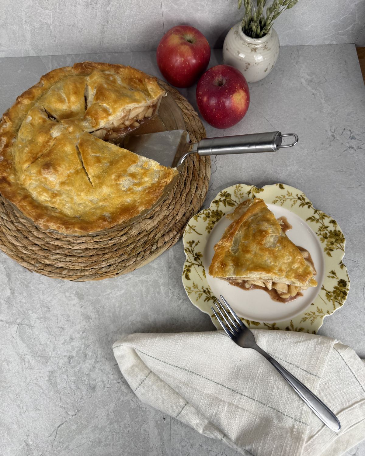 A double-crust apple pie with one slice cut and served on a floral plate, red apples and a white vase behind.