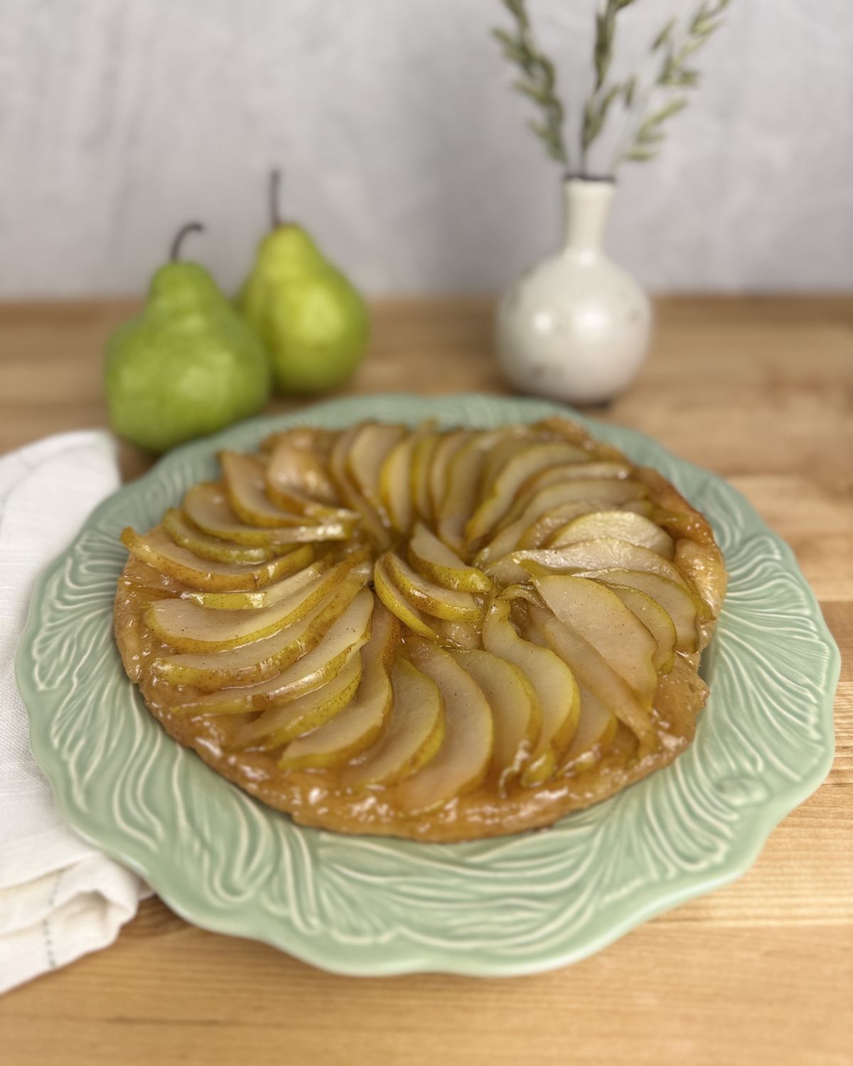 A pear tarte tatin on a green leaf-pattern plate, two green pears and a small white vase behind it.