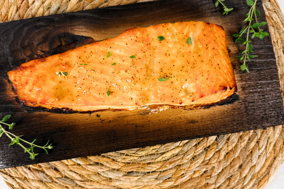 A roasted salmon fillet on a charred cedar plank set on a woven rope trivet.