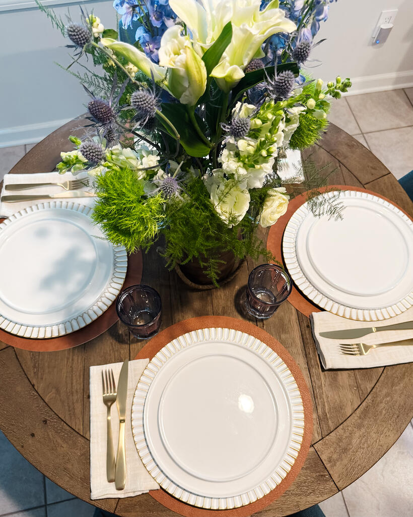 A round wooden table seen from above, set for three with white plates, wood chargers and a low arrangement of white lilies and blue delphinium.