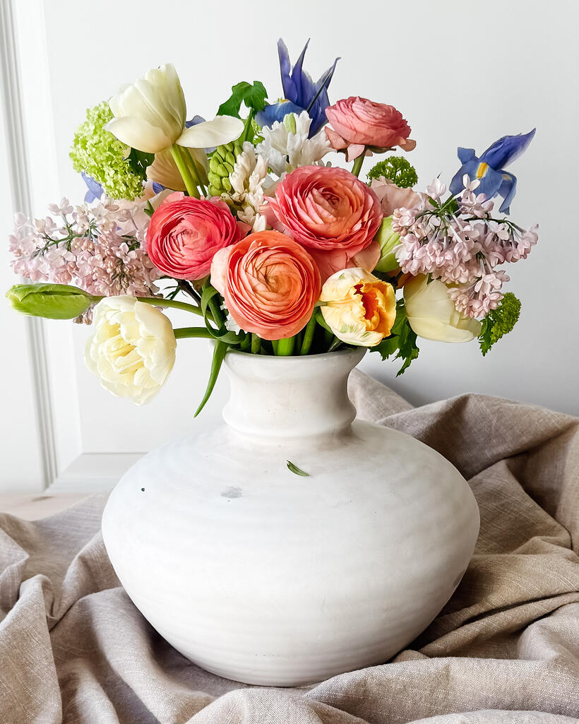 A wide white ceramic vase of ranunculus, tulips and lilac on rumpled linen.