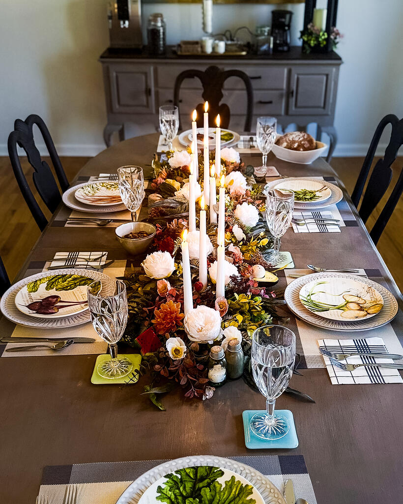 A dining room at dusk, the long table lit by tapers with a dark sideboard behind.