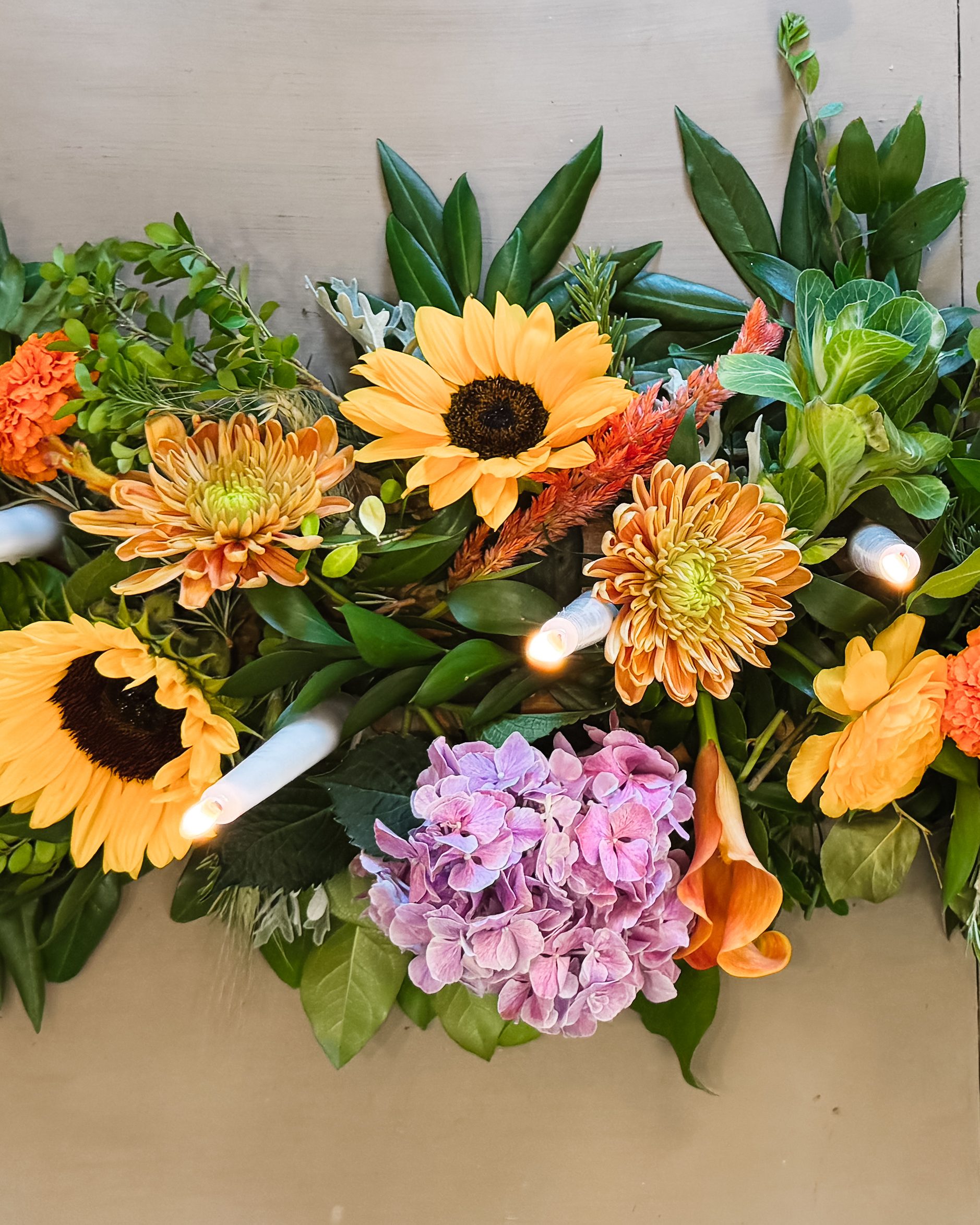 Seen from above, a runner of sunflowers, spider mums, pink hydrangea, an orange calla lily and greenery, white tapers lit among the flowers.