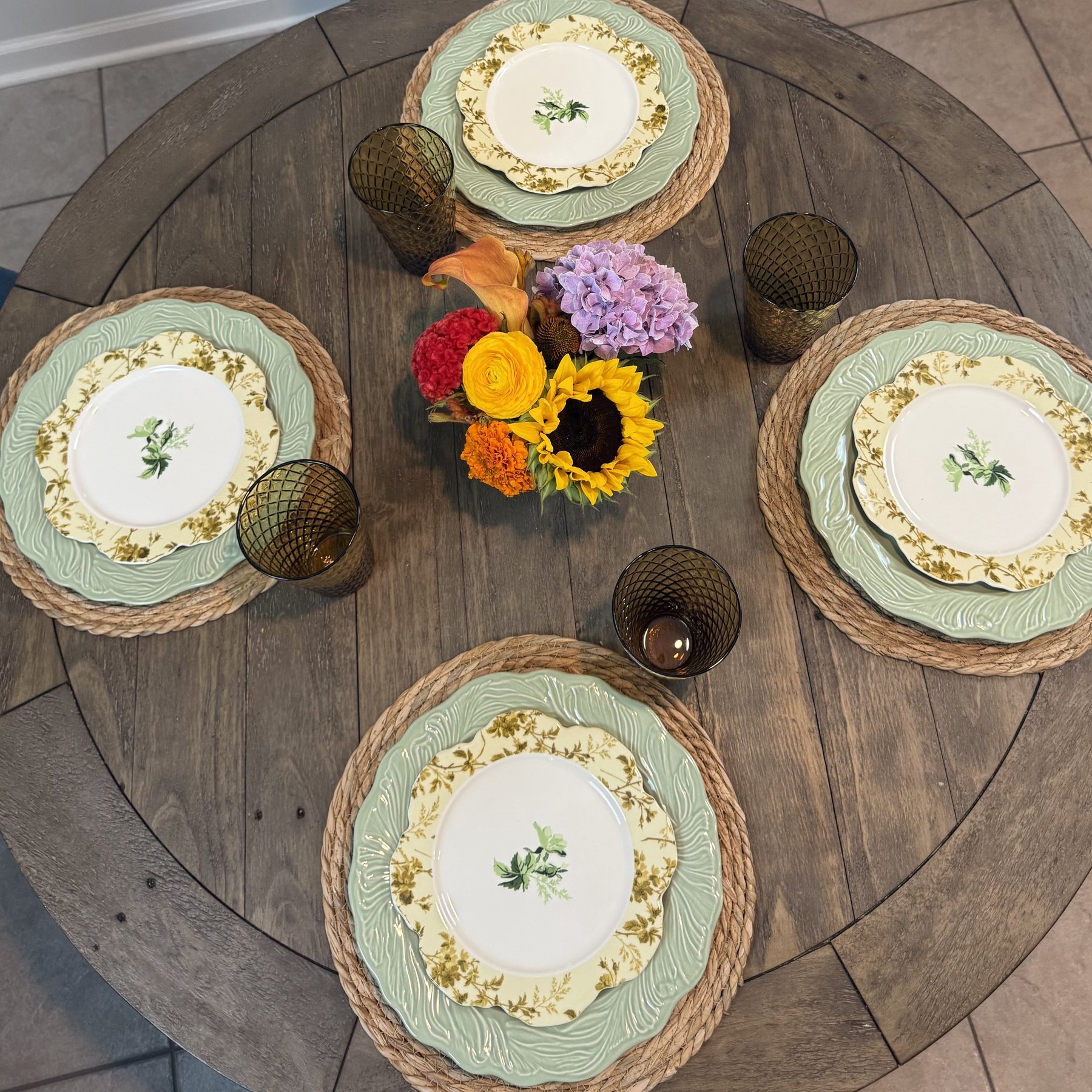 A round wooden table seen from above, set for four with floral plates on green leaf-pattern chargers and woven mats, amber glasses, and a small bunch of sunflower, hydrangea, calla lily, celosia and marigold in the middle.