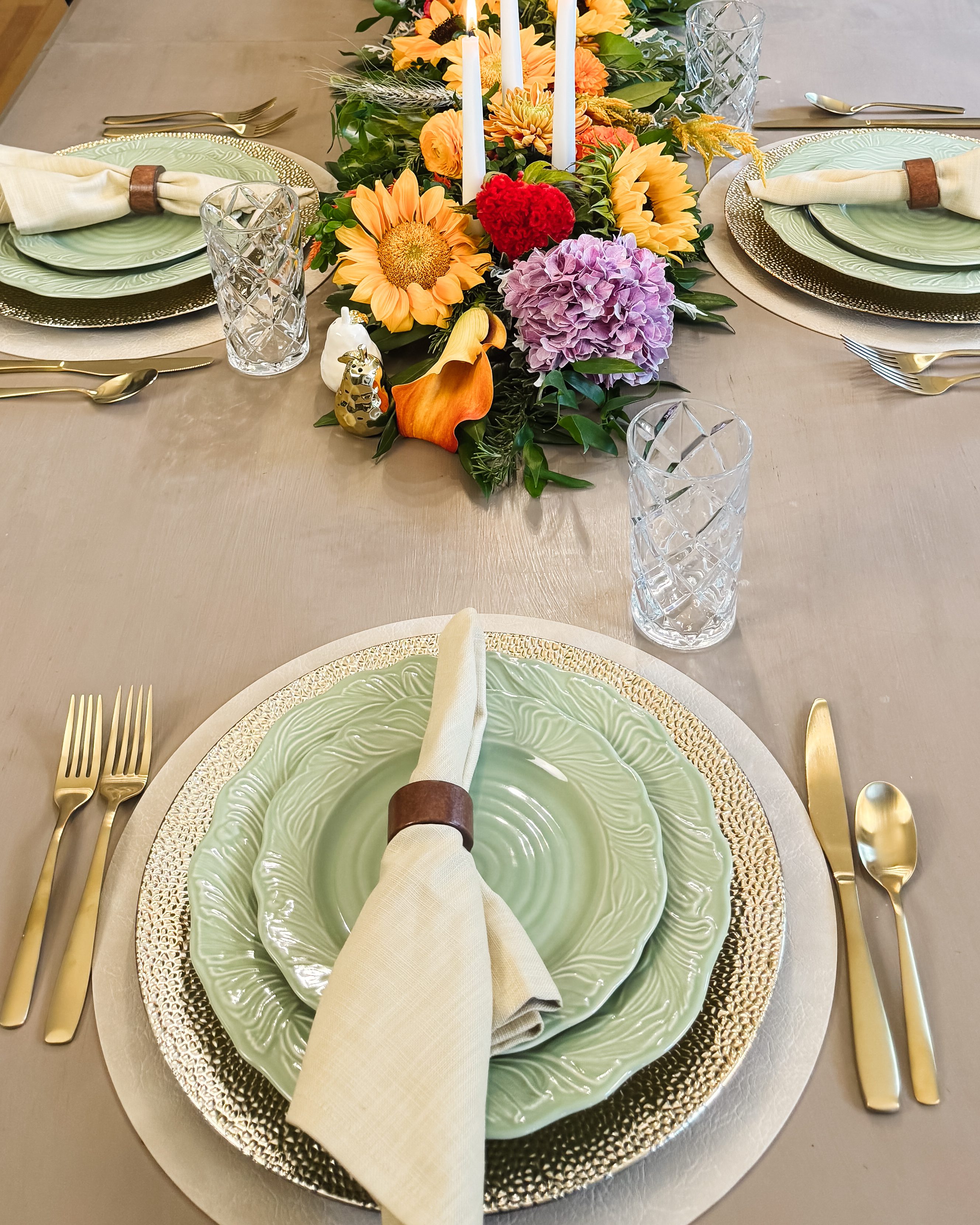 A place setting of stacked green leaf-pattern plates on a hammered silver charger, a linen napkin in a leather ring, gold flatware and a crystal glass.