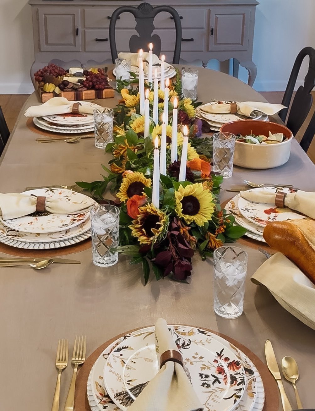 A long table set with autumn-patterned plates, linen napkins in leather rings and a runner of sunflowers and dark red foliage with white tapers.